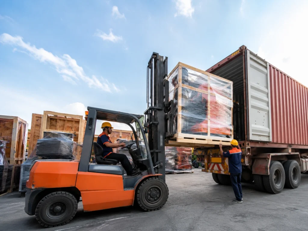 Forklift loading wrapped machinery into a shipping container in an outdoor logistics yard.